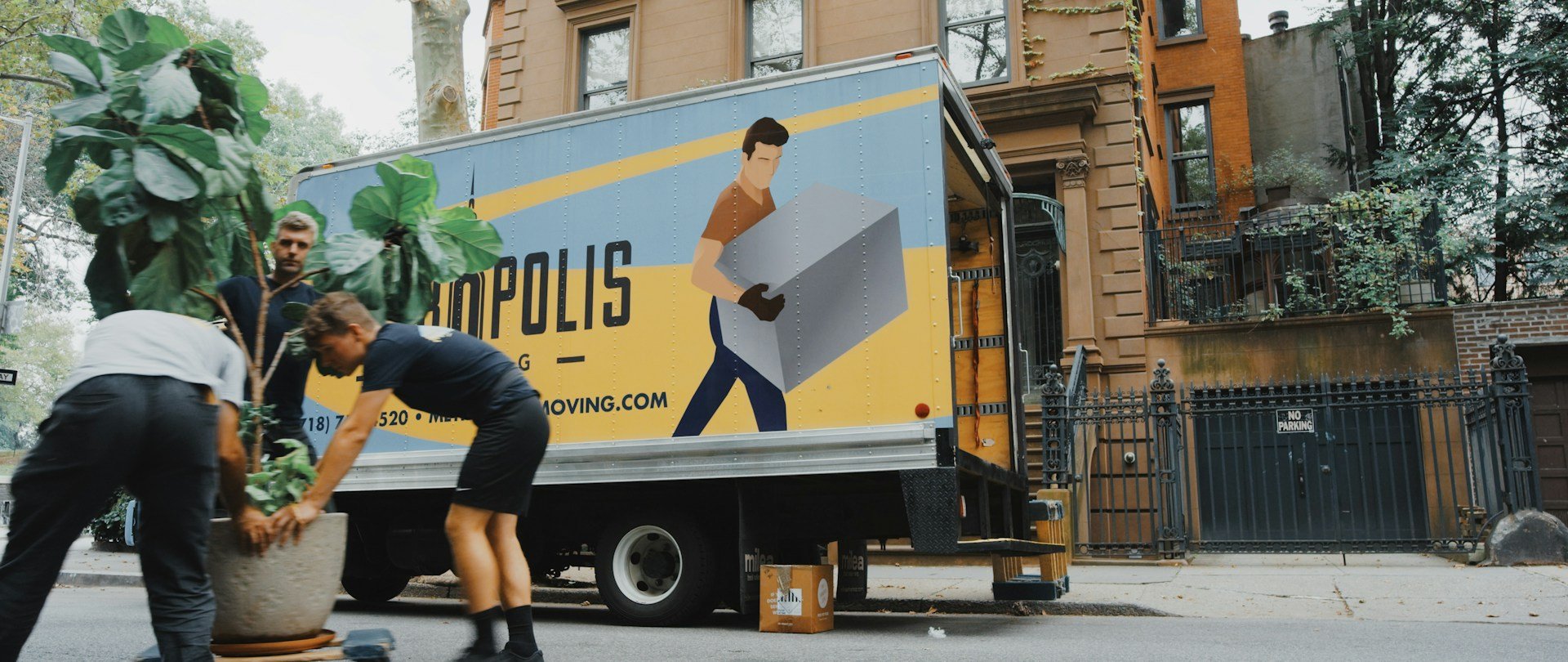 The image shows a moving truck parked on a city street, with three people standing next to it and one person bending over to pick up a plant from the ground. The truck is adorned with an advertisement for "Apolis Movers" in yellow and blue colors. In the background, there are buildings of varying heights, including a tall brown building that stands out against the sky.